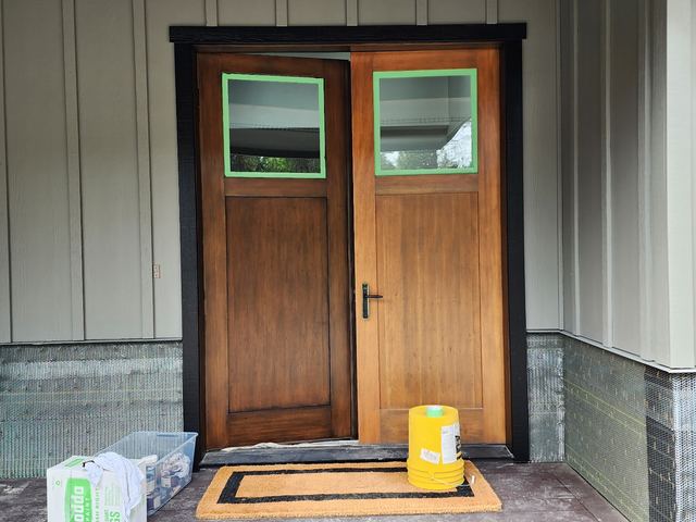 A pair of stained wood entry doors under a covered porch, their glass masked off with tape, against sage-green board-and-batten siding.
